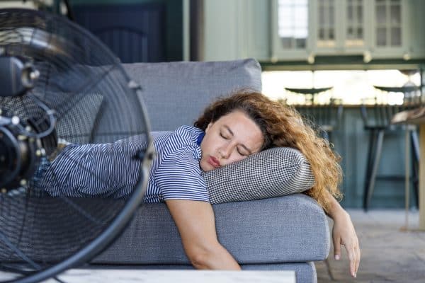 Young Woman Lying In Front Of The Electric Fan At Home, Overwhelmed By The Heat