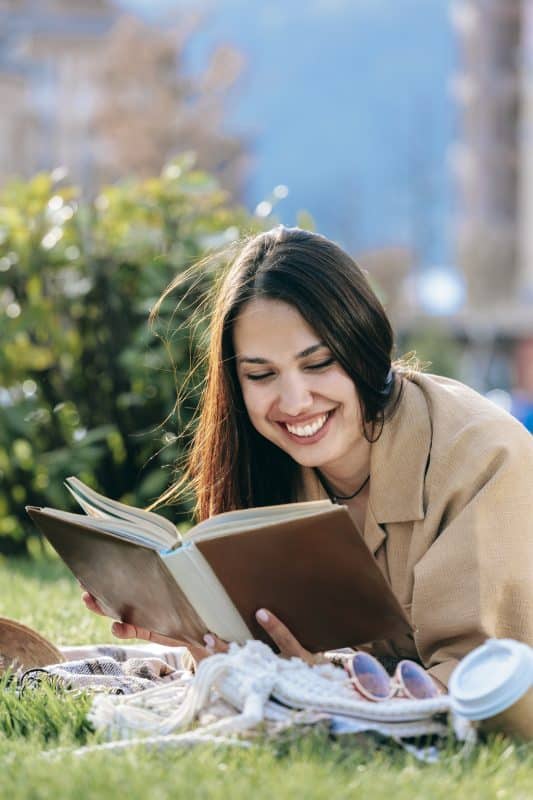 Happy Young Woman With Dark Hair Reading Book Lying At Park