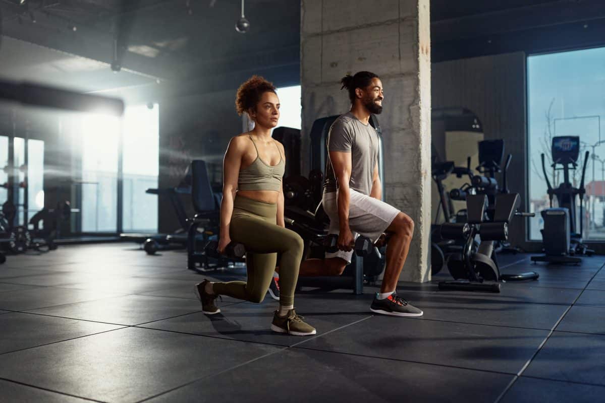 Athletic Couple Exercising Lunges With Dumbbells In A Gym.