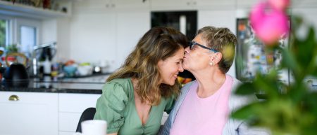 Senior Mother Kissing Adult Daughter On Forehead Indoors At Home.